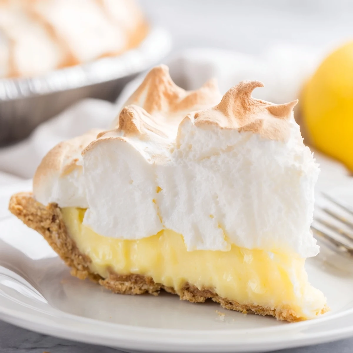 A whole Lemon Meringue Pie with Graham Cracker Crust has a fork taking a creamy slice next to fresh lemon wedges.