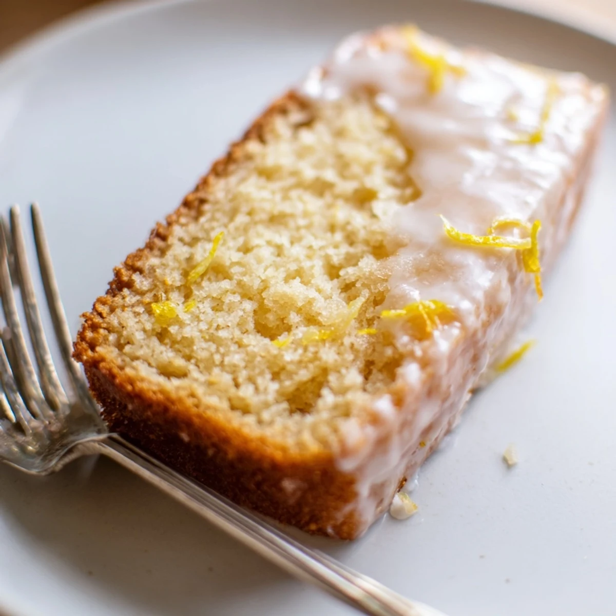 A thick slice of homemade lemon bread loaf with a shiny white glaze dripping down the sides, served on a rustic plate.