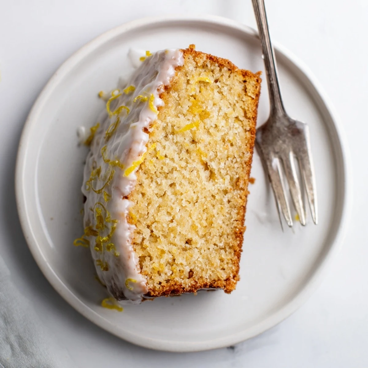 A close-up of a glazed lemon bread loaf slice on a white plate, perfect for an afternoon snack with tea.