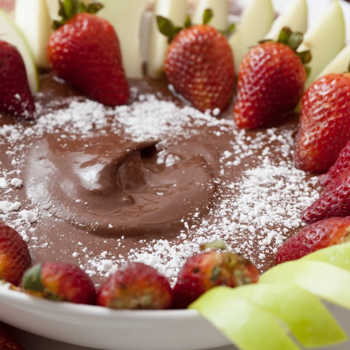 Creamy chocolate appetizer fruit dip with glossy texture beside fresh strawberries, apple slices, and grapes on a marble serving board.  