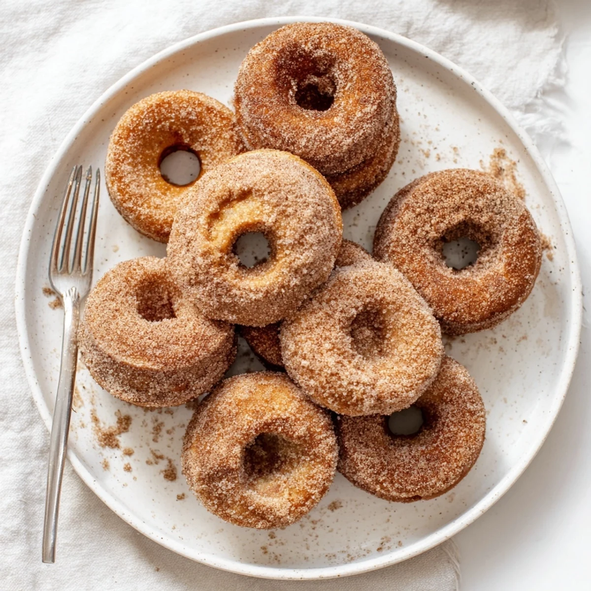 Twelve freshly baked Delicious Baked Cinnamon Sugar Donuts You Cant Resist arranged on a wire cooling rack with a small bowl of cinnamon sugar for dipping.