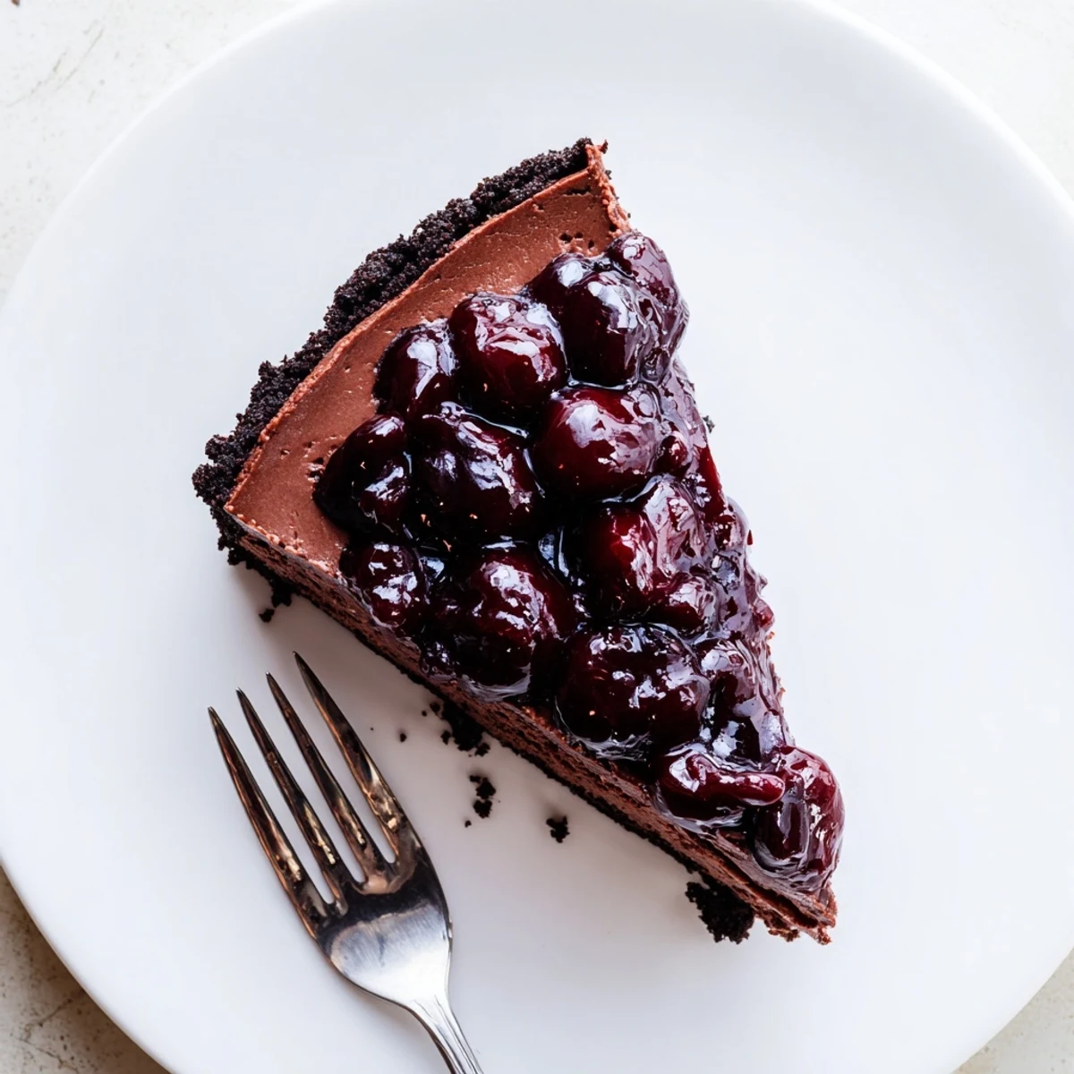 Creamy dark chocolate cherry cheesecake slice with glossy red fruit topping on white plate