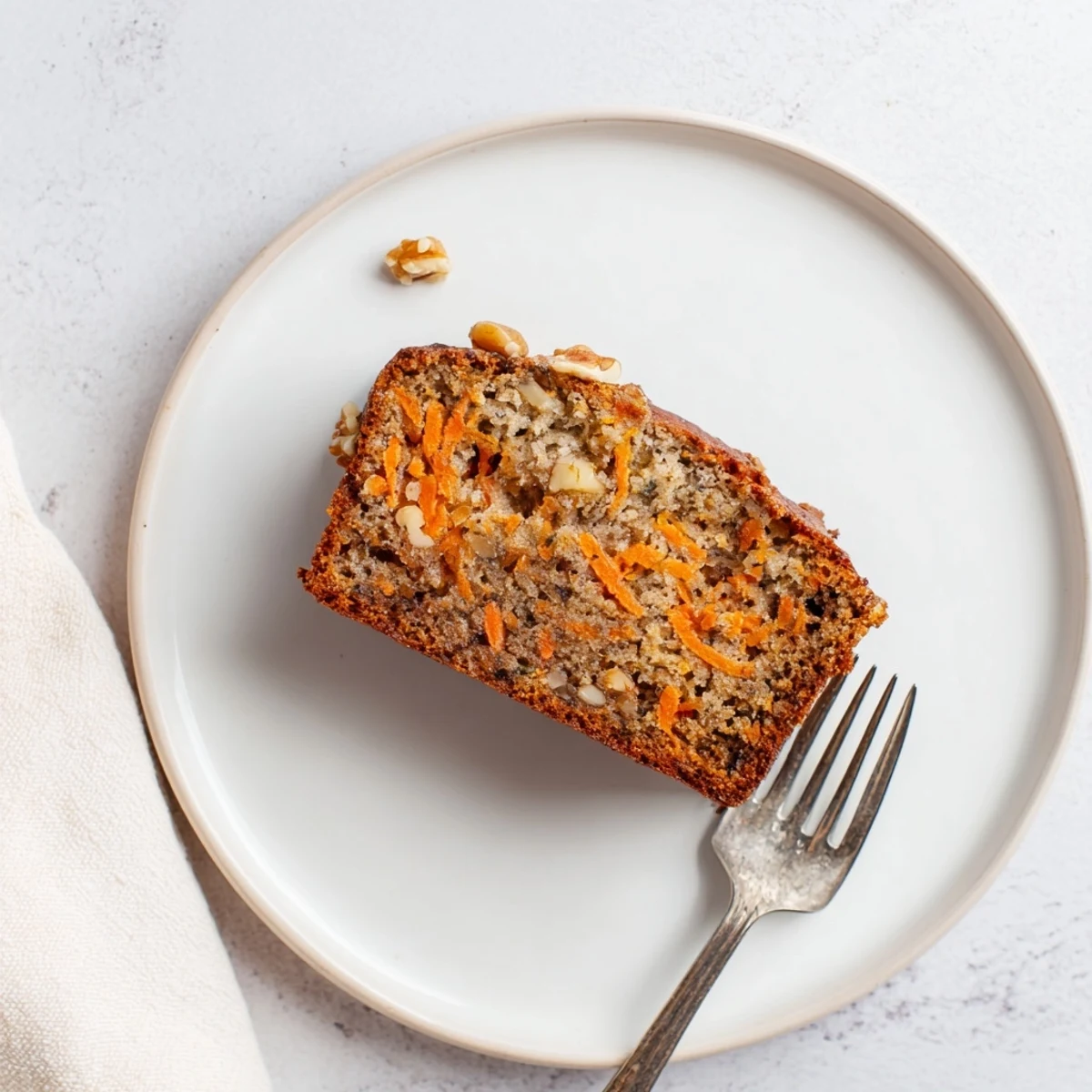 Thick slice of homemade carrot cake banana bread topped with creamy white frosting and garnished with shredded carrots and a whole walnut half