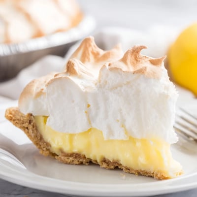 A whole Lemon Meringue Pie with Graham Cracker Crust has a fork taking a creamy slice next to fresh lemon wedges.