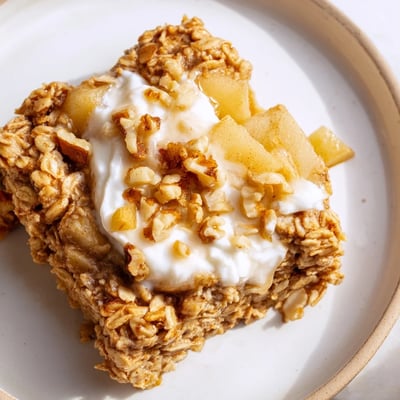 Warm Baked Apple Oatmeal With Greek Yogurt sits on a white plate, revealing tender apples inside the golden oats.