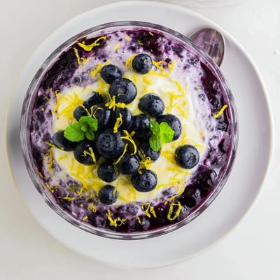 The dessert features soft peaks of whipped cream folded with lemon curd, topped with juicy blueberry sauce and zest.